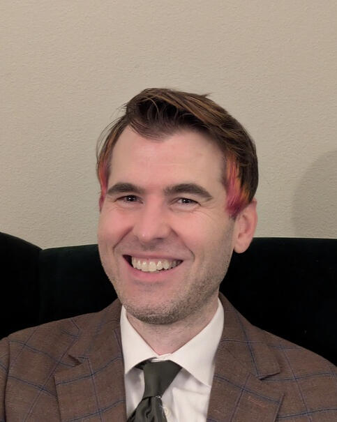 Headshot of Trevor Murphy. A white man with a five o'clock shadow, a big smile, and short brown hair dyed pink and orange on the sides. He is wearing a brown windowpane suit and green tie, seated in a green velvet chair, in front of a cream-colored orange-